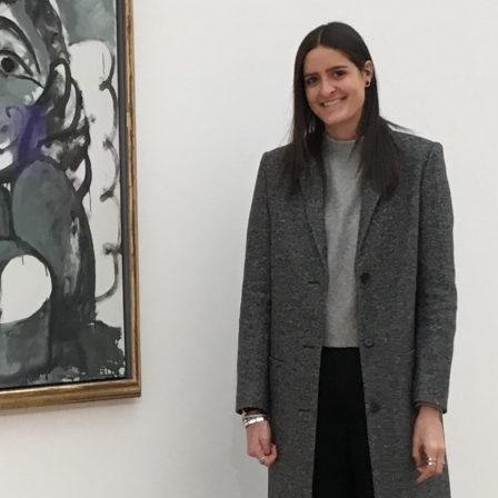 Image of Claire Renaud. Claire in standing in front of a painting that is obscured from view. She has medium length black hair and is wearing a long gray coat over a lighter gray turtleneck.